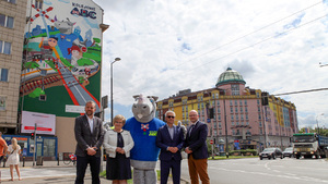 Cztery osoby wraz z maskotką Rogatkiem na tle muralu Kolejowego ABC na Placu Zawiszy w Warszawie.