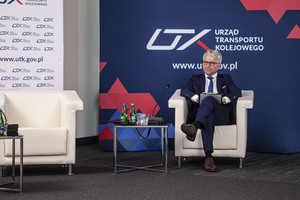 Prezes UTK dr inż. Ignacy Góra podczas konferencji z okazji 18 lat Urzędu Transportu Kolejowego. Prezes siedzi na fotelu przed nim stolik na nim woda do picia za Prezesem granatowa ścianka z logo UTK