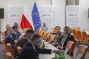 Wnętrze sali konferencyjnej z rozłożonymi stołami przy nich siedzą osoby pośrodku zdjęcia flaga Polski i Unii Europejskiej