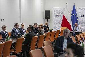 Wnętrze sali konferencyjnej z rozłożonymi stołami przy nich siedzą osoby po prawej stronie zdjęcia flaga Polski i Unii Europejskiej