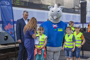 Po lewej stronie Wiceprezes UTK Marcin Trela po środku zdjęcia dzieci ubrane w jasnozielone kamizeli kampanii Kolejowe ABC a za nimi maskotka Rogatek
