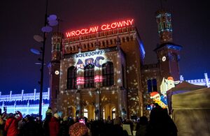 Zdjęcie dworca Wrocław Główny oświetlonego kolorowymi iluminacjami. Na elewacji logo Kolejowe ABC. Na dachu budynku neon Wrocław Główny.