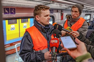 Mężczyzna - inspektor UTK - w szarej kurtce i pomarańczowej kamizelce odblaskowej stoi przed dziennikarzami w środku składu pociągu i tłumaczy przebieg kontroli. Przed nim mikrofony. Za nim okno pociągu. Mężczyzna ma krótkie włosy.
