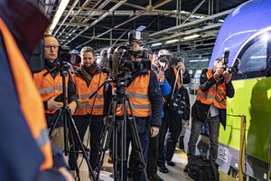 Widok dziennikarzy zebranych podczas konferencji. Dziennikarze ubrani są w pomarańczowe kamizelki odblaskowe robią zdjęcia przed dziennikarzami ustawione są kamery. Osoby zgromadzone są w hali serwisowej pociągów.