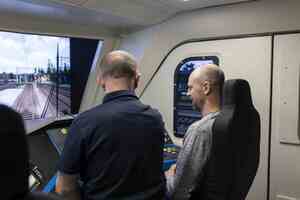 Wnętrze symulatora. Na krześle siedzi mężczyzna przed nim panel sterowania oraz ekran na którym wyświetlana jest grafika symulująca przejazd lokomotywą.