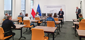 Zdjęcie sali egzaminacyjnej z pojedynczymi stolikami i fotelami. Uczestnicy spotkania siedzą i słuchają wypowiedzi Prezesa UTK. W tle widoczna flaga Polski i Unii Europejskiej.