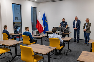 Trzech mężczyzn siedzi przy stołach w sali egzaminacyjnej, trzech mężczyzn stoi i tłumaczy zasady egzaminu. Widoczna flaga Polski i Unii Europejskiej.